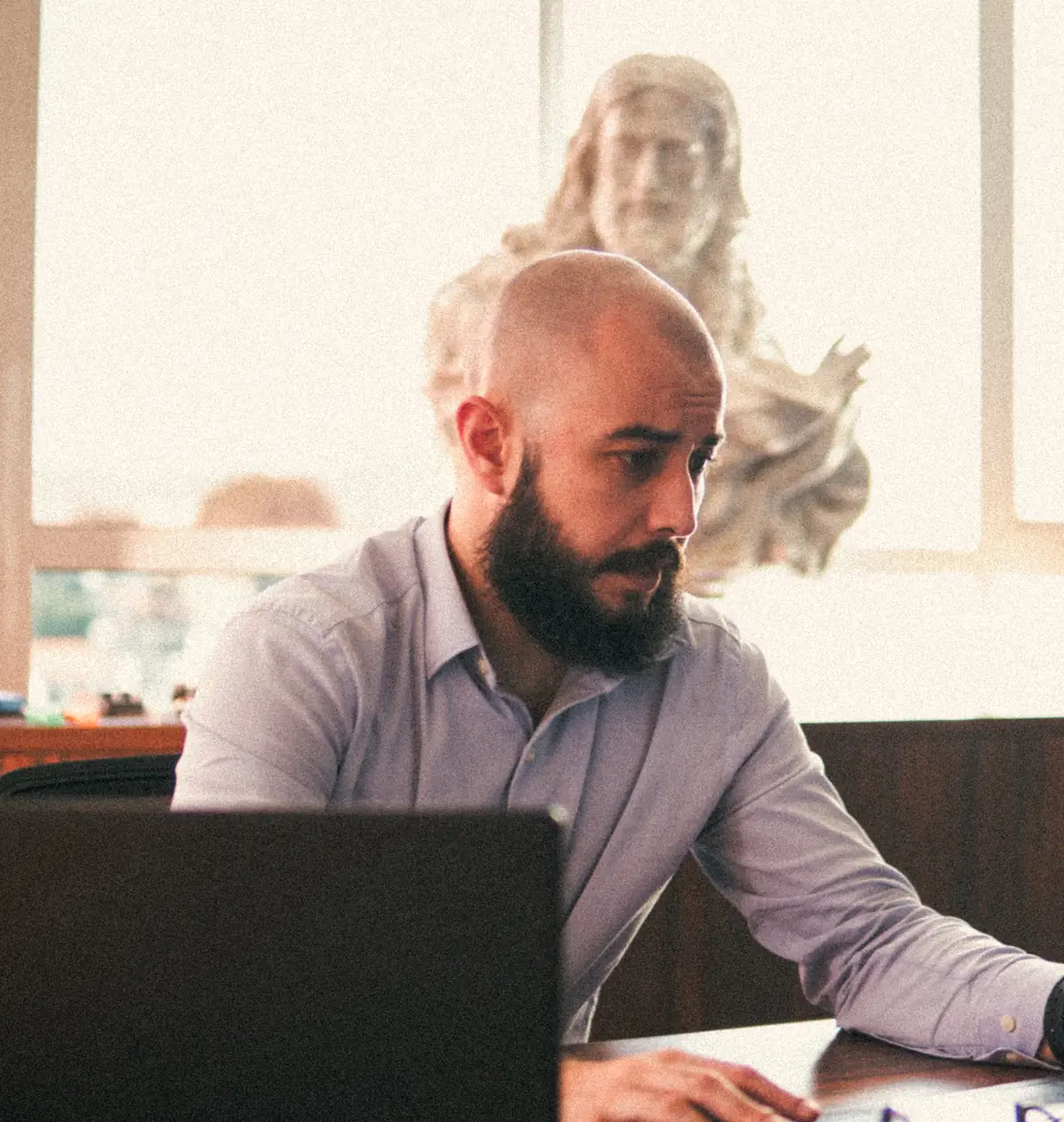 Artur Vargas concentrado ao notebook, diante de uma janela clara com um busto de mármore ao fundo.