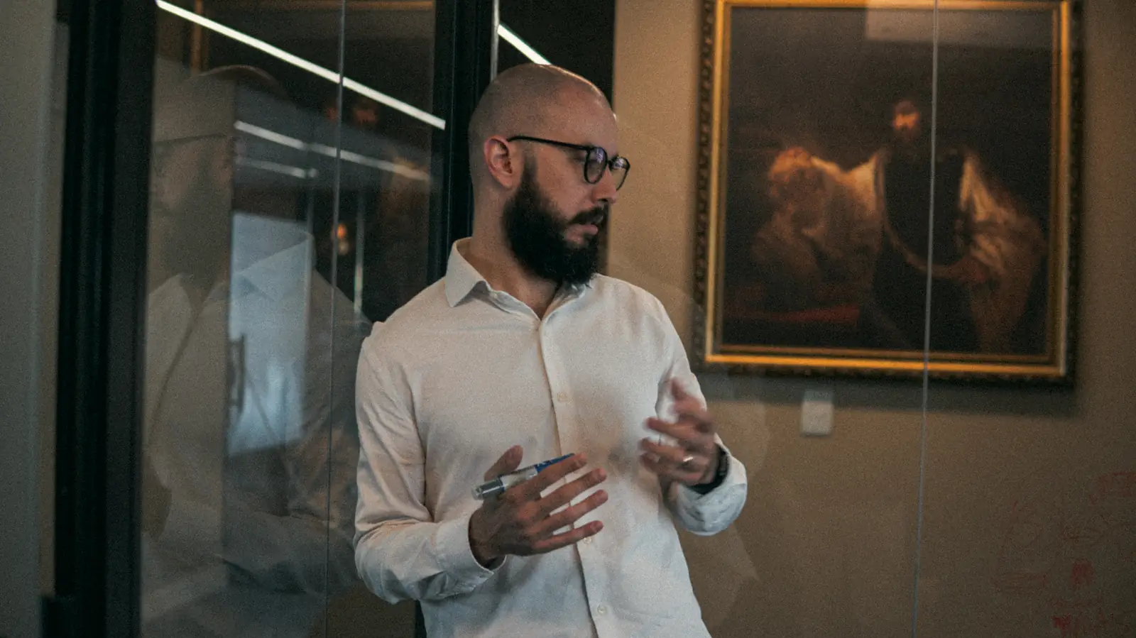 Artur Vargas, de camisa branca e óculos, explicando algo diante de uma pintura a óleo.